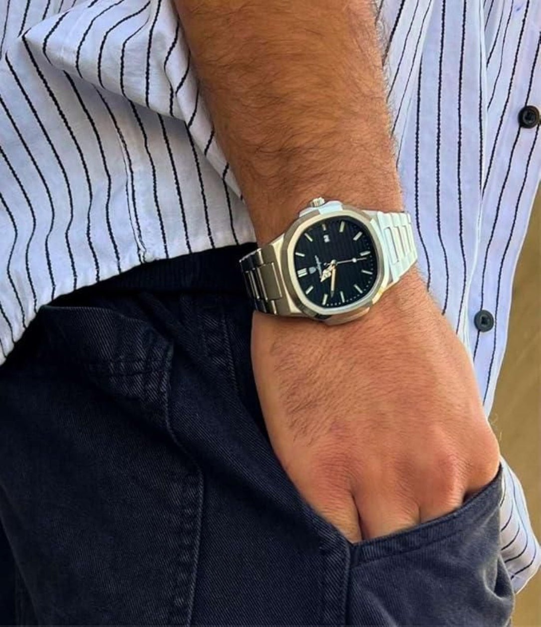 Person wearing a silver watch with a black face on a striped shirt and dark jeans.