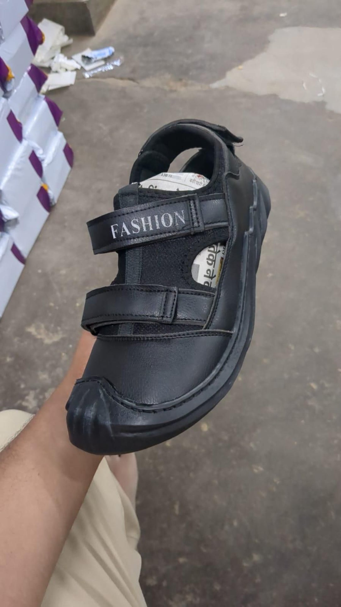 Black sandal with 'FASHION' label held by a person on a concrete surface.