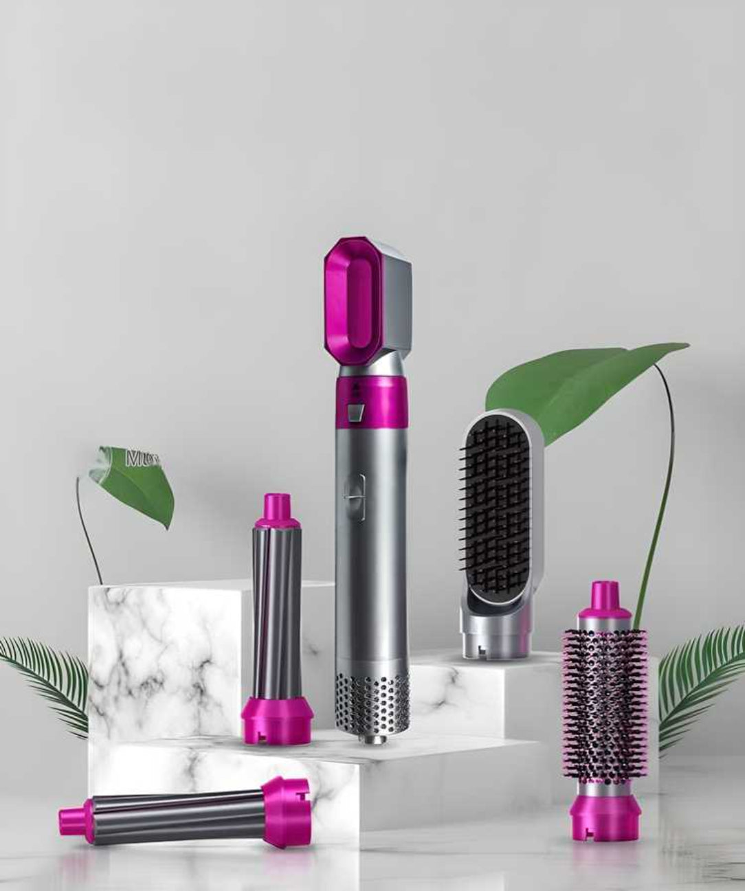Set of hair styling tools with pink and silver accents on a marble surface with green leaves.