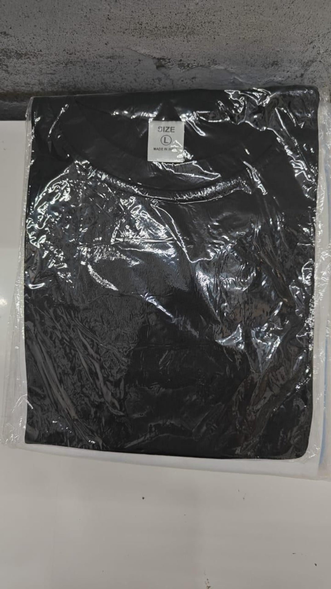 Black t-shirt with a brand logo in clear plastic packaging on a white surface.