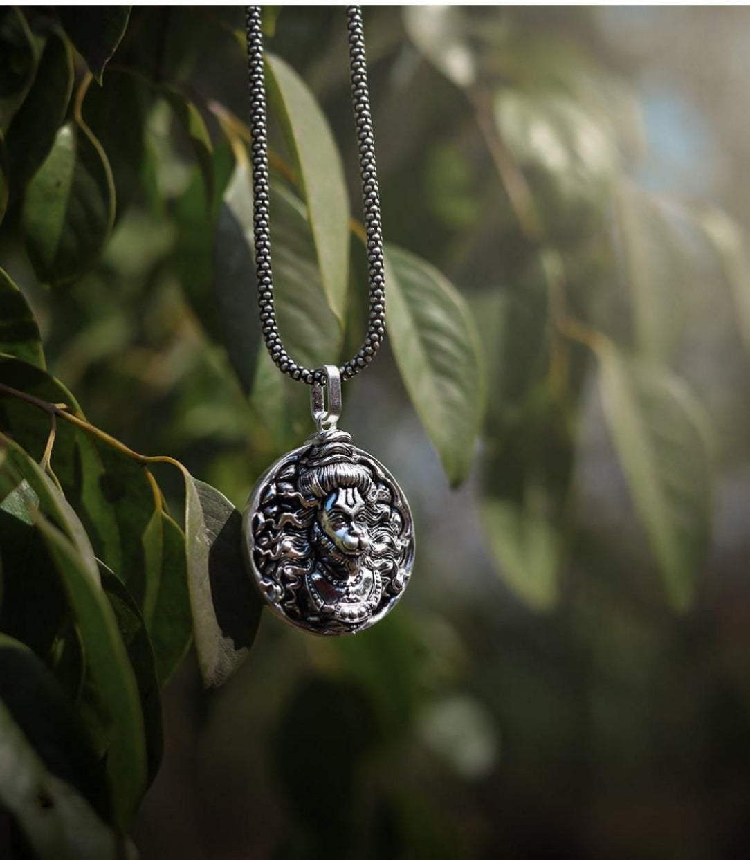 A silver chain necklace with a Hanuman pendant hanging among green leaves.