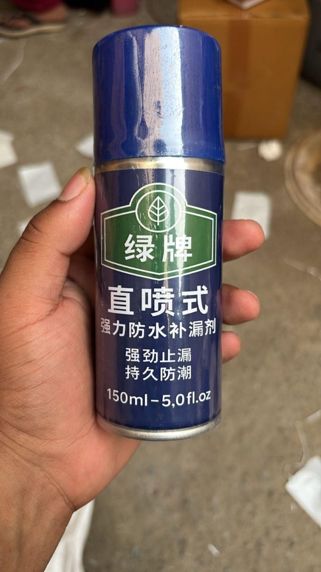 Hand holding a blue spray can labeled 'Green Brand' against a blurred background