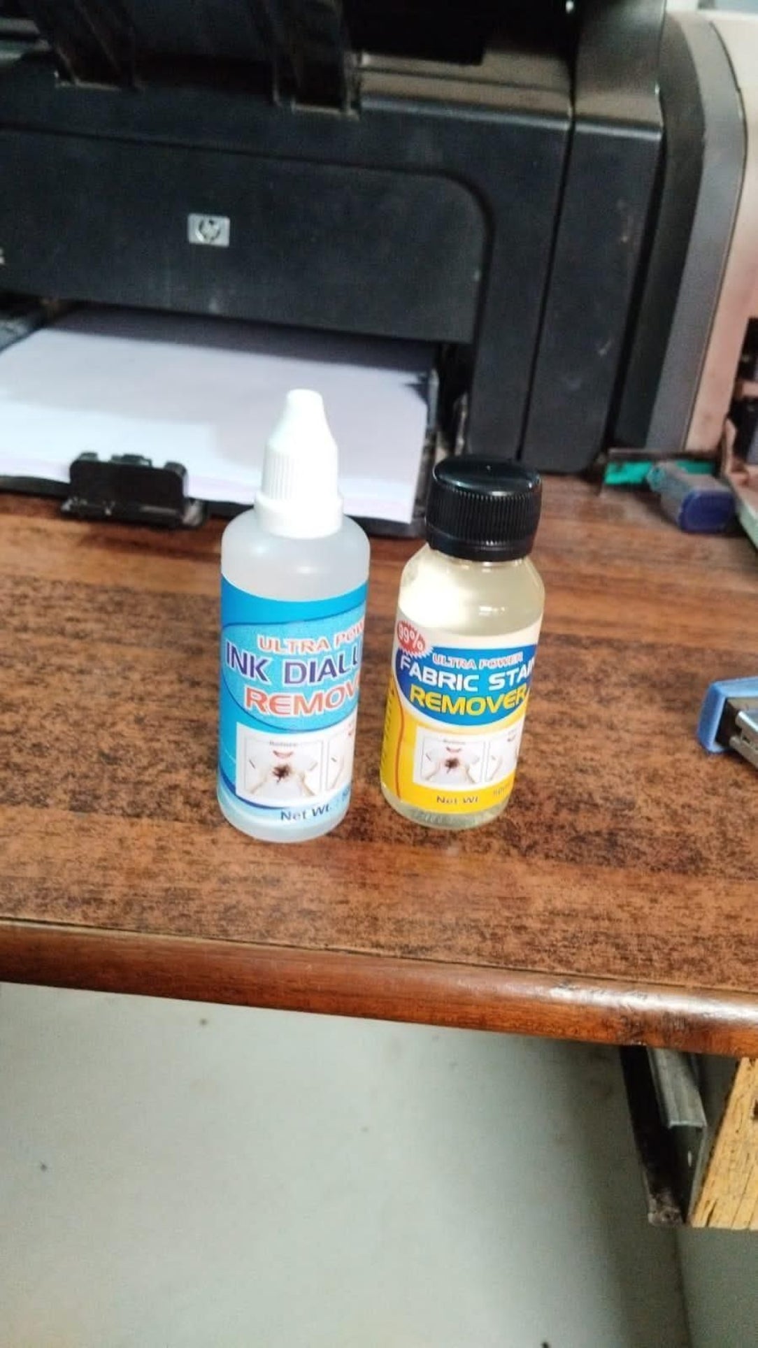 Two bottles of ink and toner cleaning solutions on a wooden surface with a printer in the background.
