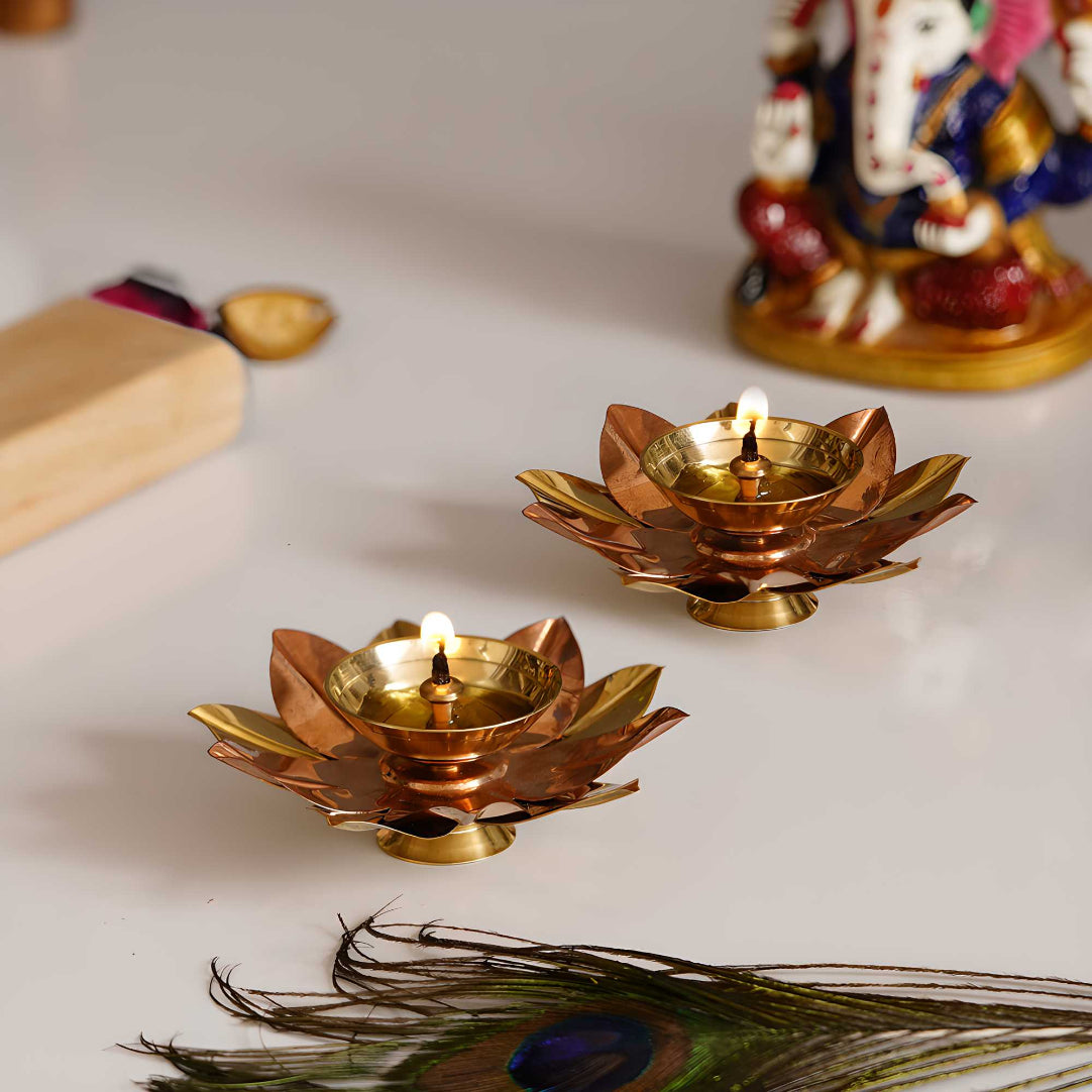 Set of two copper-colored metal diyas with a floral shape on a white surface, with a peacock feather nearby.