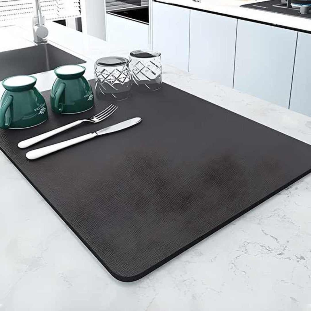 A black quick-drying mat placed on a kitchen countertop with a set of cutlery on top of it, including a green cup and a clear glass.