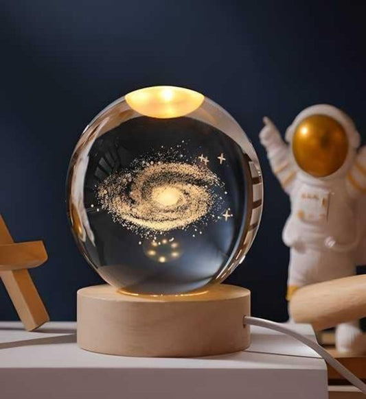 A crystal-clear 3D engraved lamp ball with a warm glow, placed on a wooden stand, with a space-themed design visible inside the ball.