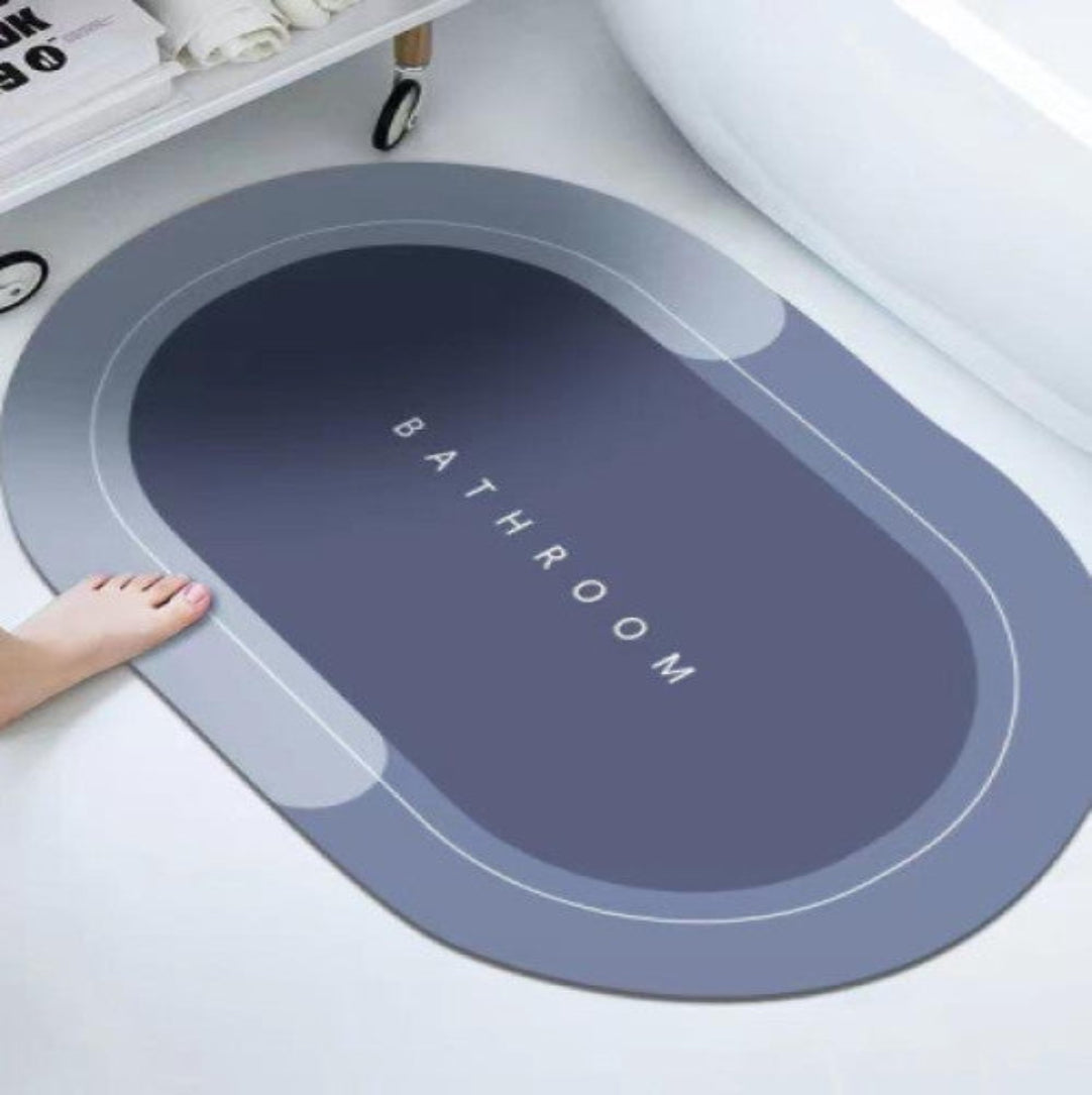 A gray bath floor mat with the word 'BATHROOM' written on it, placed on a tiled bathroom floor next to a bathtub.