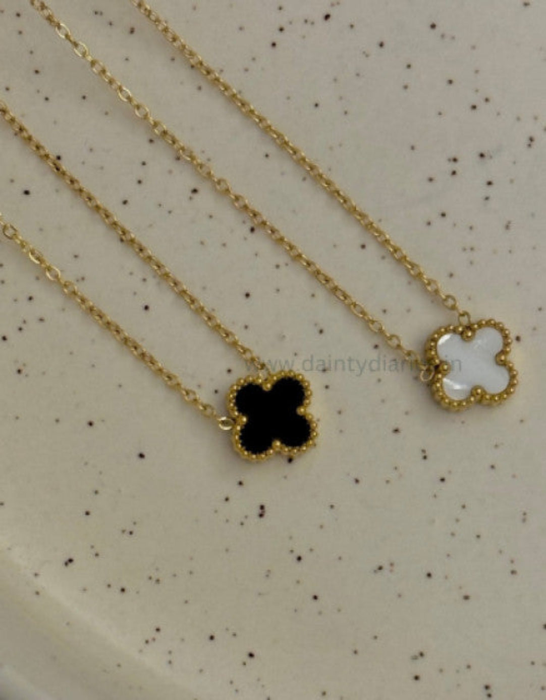 Two gold necklaces with clover pendants on a speckled beige surface.
