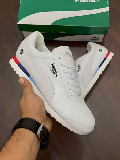 A pair of men's casual white shoes with red, blue, and black accents, held in a person's hand, with a Puma box visible in the background.