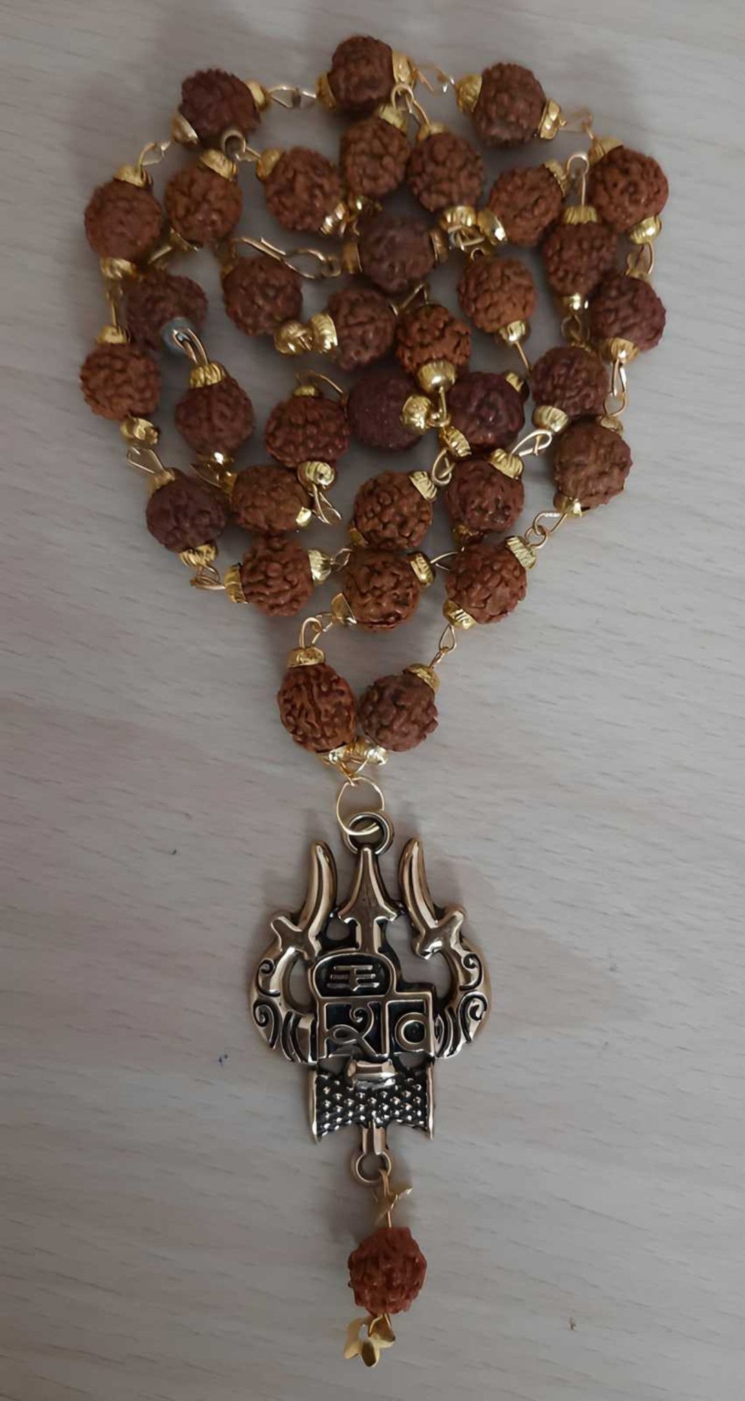 A gold-plated Rudraksha chain with intricate beads and a decorative metal hook, against a beige background.