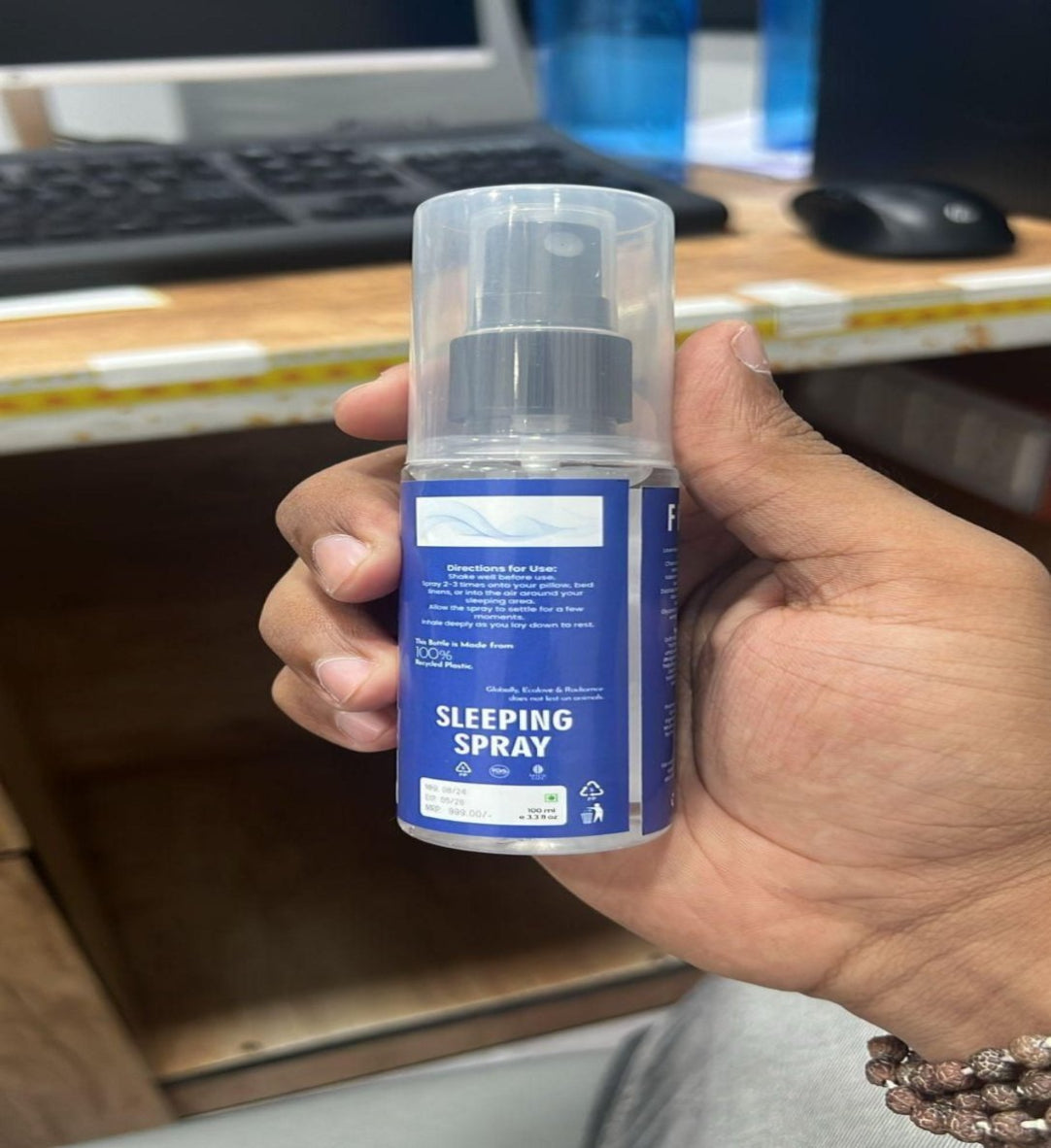 Hand holding a bottle labeled 'Sleeping Spray' in front of a computer desk.