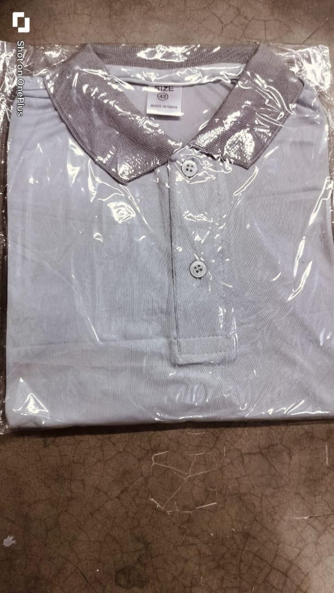 Light gray polo shirt with a collar in clear plastic packaging on a brown surface.