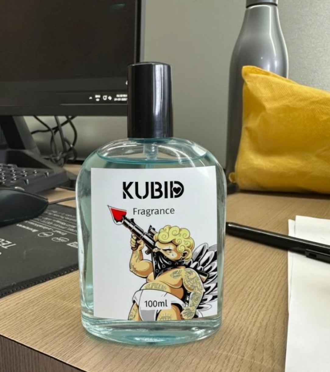 KUBID fragrance bottle on a desk with a computer and yellow pillow in the background