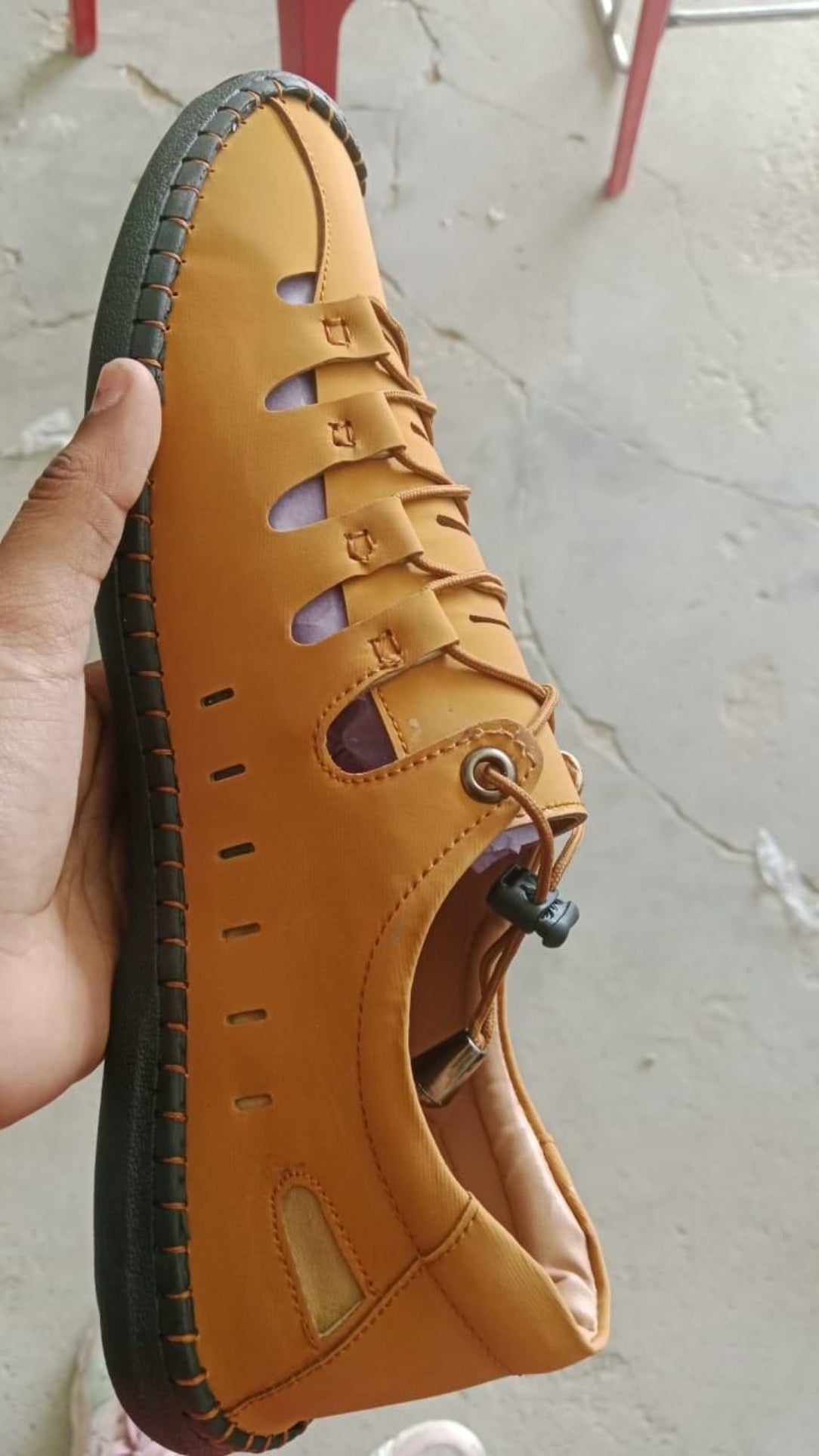 Yellow sandal with brown sole held by a hand on a light gray floor.