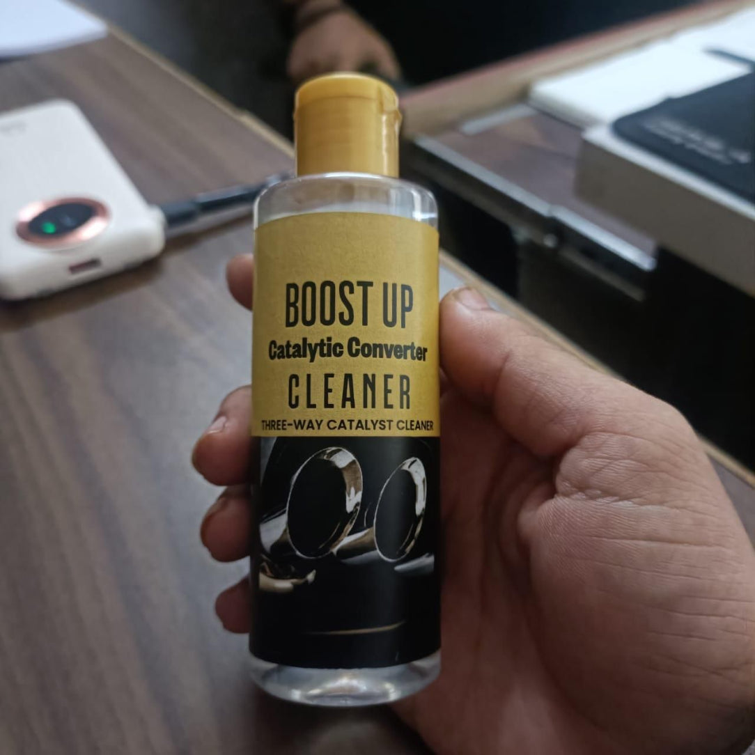 Hand holding a bottle of 'Boost Up Catalytic Converter Cleaner' on a desk.