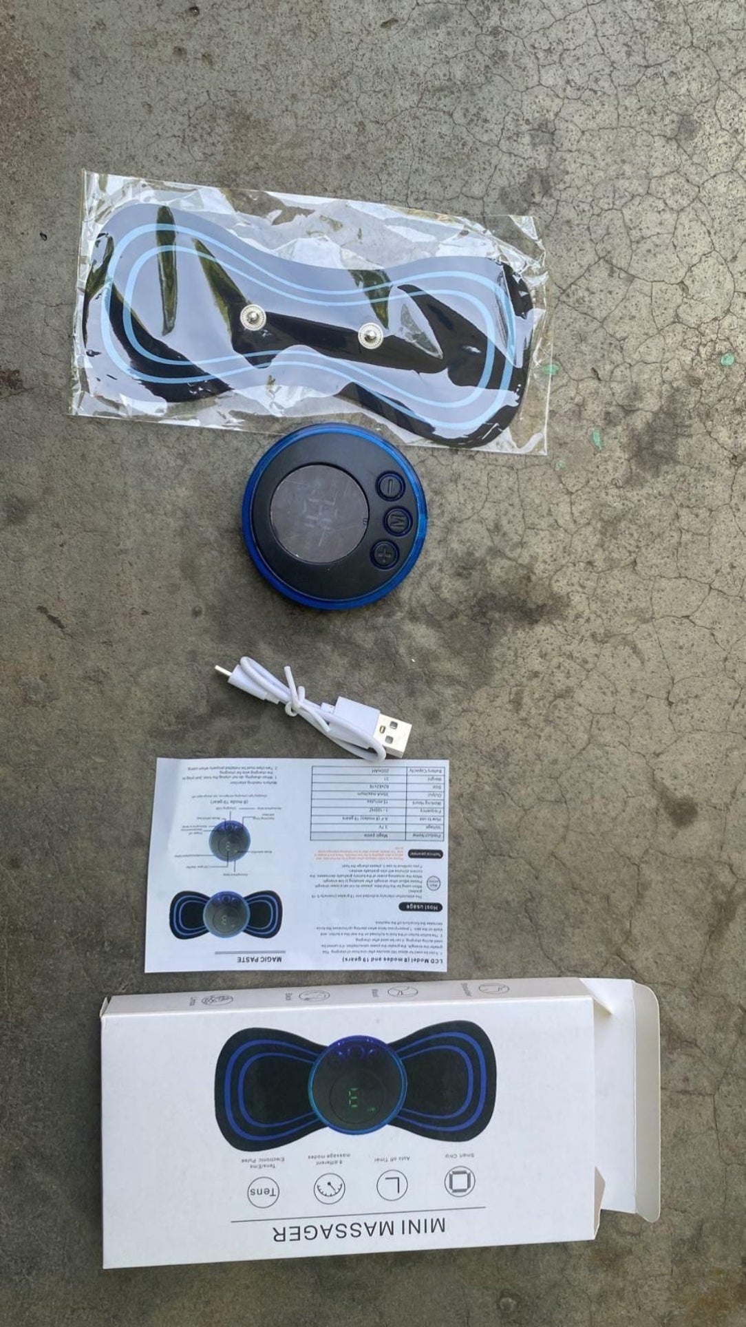 Mini massager with packaging, USB cable, and user manual on a concrete surface
