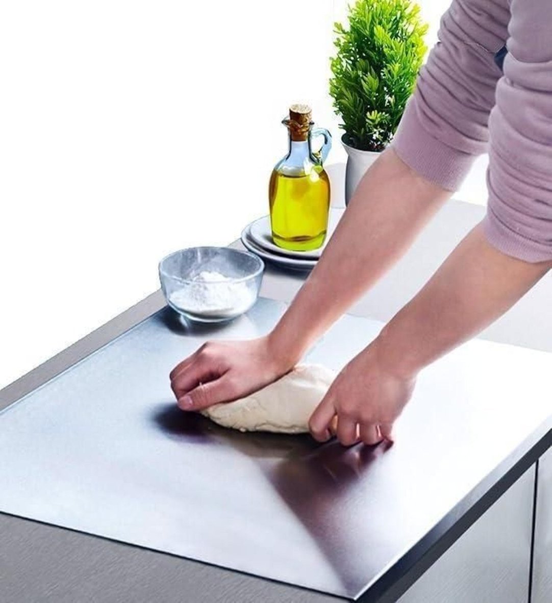 A person is using a stainless steel chopping board to cut dough, which is placed on a kitchen countertop. In the background, there is a small bowl and a potted plant, indicating a home kitchen setting.