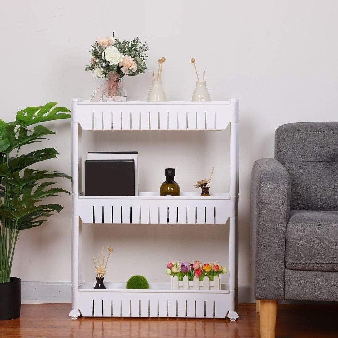 A white plastic 3-layer storage organizer rack with wheels, placed in a room next to a gray chair and decorative plants, displaying various household items on each shelf.