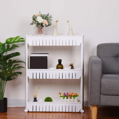 A white plastic 3-layer storage organizer rack with wheels, placed in a room next to a gray chair and decorative plants, displaying various household items on each shelf.