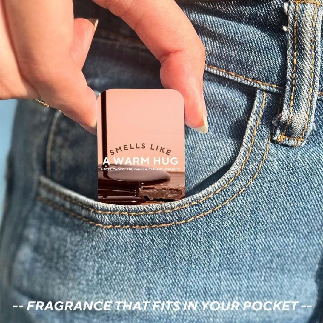 A solid perfume stick with the phrase 'Smells like a warm hug' printed on it, held between fingers with a denim background.