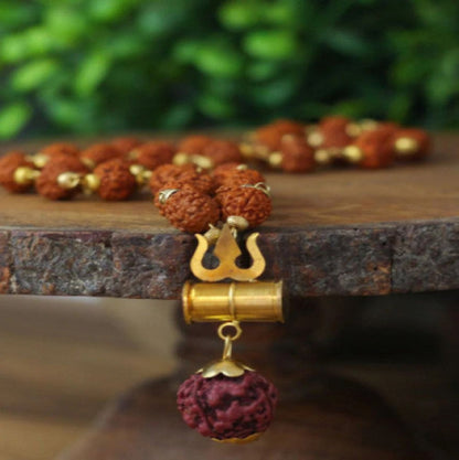 A gold-plated Trishul Rudraksha Mala with brown beads and a gold-colored metal Guru bead, displayed on a wooden surface.
