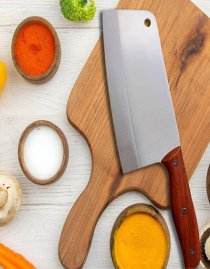 Wooden cutting board with a large knife on a white surface with various spices and vegetables.